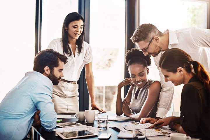 pricing-program Five people in a meeting room are gathered around a table, engaged in discussion, with documents and a laptop in front of them.